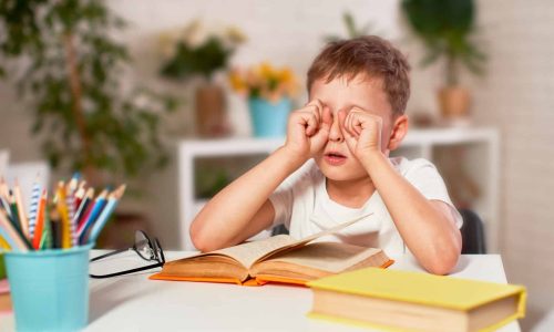 the child is tired of learning. home schooling, homework. the boy rubs his eyes from fatigue reading books and textbooks. a little boy student sitting at a table with books. vision problems