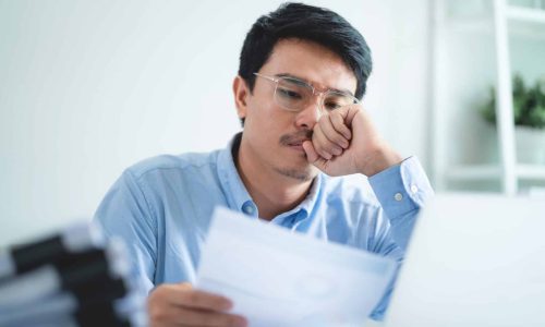 A man is sitting at a desk with a laptop and a stack of papers. He is looking at a piece of paper and he is in a state of deep thought
