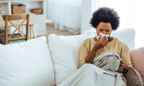 Shot of a mature woman blowing her nose with a tissue at home. Woman sneezing in a tissue in the living room. Worried African American woman sitting at home while being sick